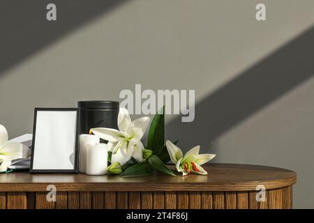 Cadre funéraire vierge, urne mortuaire, bougies allumées et fleurs de lys sur une table en bois près du mur gris Banque D'Images