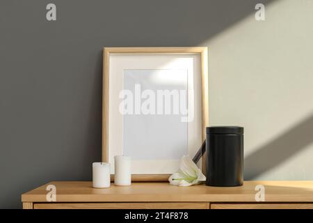 Cadre funéraire vierge, urne mortuaire, bougies et fleur de lys sur armoire en bois près du mur gris Banque D'Images