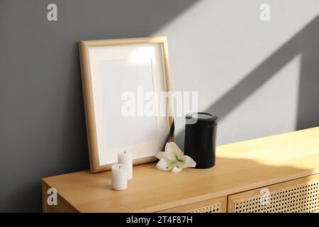 Cadre funéraire vierge, urne mortuaire, bougies et fleur de lys sur armoire en bois près du mur gris Banque D'Images