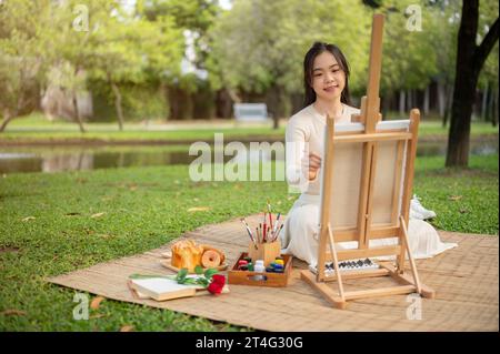 Une jeune femme asiatique belle et créative aime peindre sur une toile sur un chevalet dans un beau parc. Concept de loisirs de créativité Banque D'Images