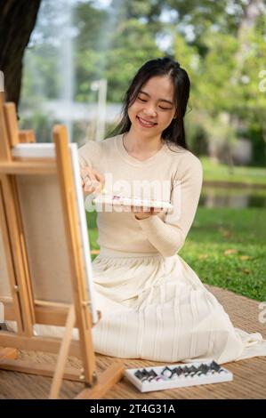 Une femme asiatique heureuse et jolie aime peindre sur une toile sur un chevalet dans un parc verdoyant le week-end. concept d'activité de créativité Banque D'Images