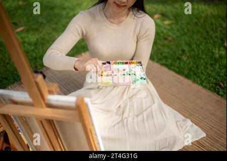 Image en gros plan d'une jeune artiste asiatique talentueuse et créative mélange la couleur de l'eau sur une palette, peignant sur une toile dans un parc vert. creativi Banque D'Images