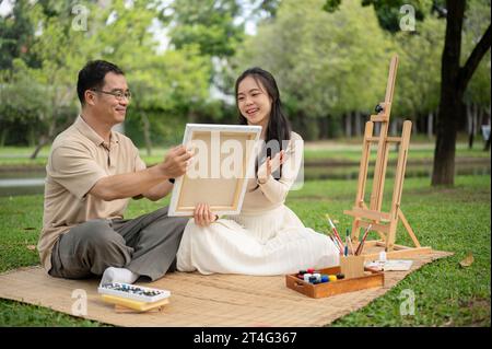 Une fille asiatique charmante et joyeuse aime pique-niquer dans un parc verdoyant avec son père, passer du temps à créer et peindre une image sur une toile ensemble. HAP Banque D'Images