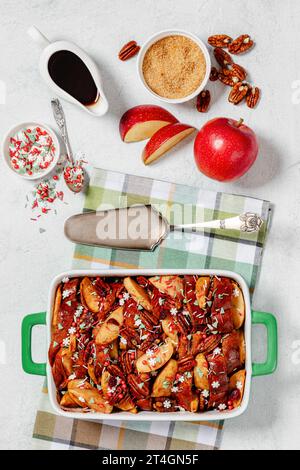 Pain grillé français aux pommes au four recouvert de noix de pécan et de saupoudrages de sucre de noël dans un plat allant au four avec des ingrédients et une pelle à gâteau sur une table en béton blanc, Banque D'Images