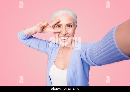 Une dame âgée fait un selfie sur l'appareil photo d'un smartphone faisant un v-sign près des yeux isolés sur fond violet clair avec un espace de copie pour le texte Banque D'Images