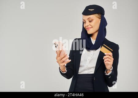 hôtesse souriante dans l'uniforme des compagnies aériennes arabes tenant la carte de crédit et regardant smartphone sur gris Banque D'Images