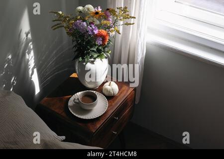 Intérieur de chambre Moody. Tasse de café, citrouille blanche sur table de chevet en bois. Vase en céramique au soleil. Bouquet de dahlia, cosmos, fleurs de solidago Banque D'Images