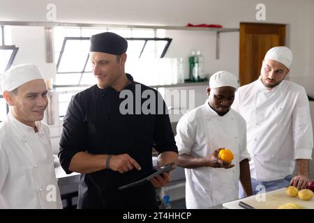 Professeur de chef diversifié utilisant la tablette numérique et enseignant la cuisine aux étudiants masculins en cuisine Banque D'Images