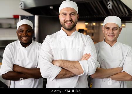 Portrait de divers chefs masculins souriants avec les bras croisés debout côte à côte dans la cuisine commerciale Banque D'Images