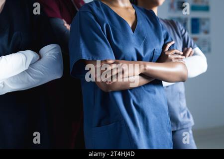Section médiane de divers médecins masculins et féminins portant des gommages croisant les bras dans le couloir à l'hôpital Banque D'Images