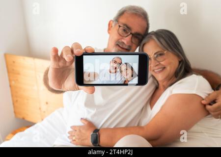 Heureux vieux mari et femme européens faire selfie sur l'écran du téléphone sur le lit dans la chambre Banque D'Images