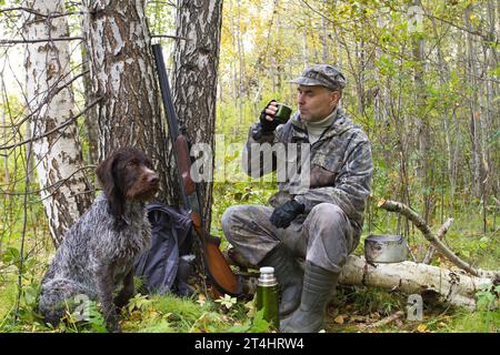 un chasseur est assis sur un arbre tombé et boit du thé dans un thermos, son chien est assis à côté de lui Banque D'Images