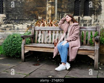 Jeune femme avec les cheveux bruns de longueur d'épaule assis sur un banc un jour d'automne à Deans Park à York Yorkshire Angleterre Banque D'Images