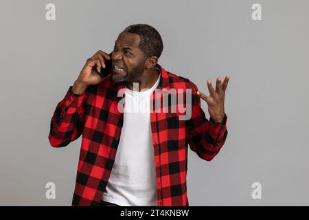 Homme barbu noir agressif en colère portant une chemise à carreaux rouge criant tout en parlant au téléphone Banque D'Images
