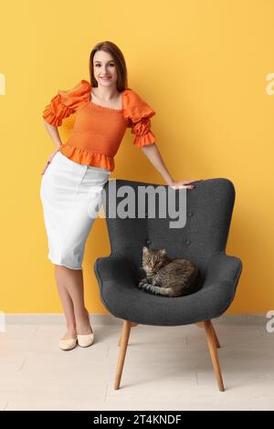 Jolie jeune femme avec chat tabby mignon sur fauteuil sur fond orange Banque D'Images