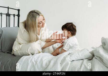 Portrait d'une petite fille et d'une mère qui sont assises sur le lit le matin après le sommeil. Une mère heureuse tend les mains à sa fille et à smi Banque D'Images