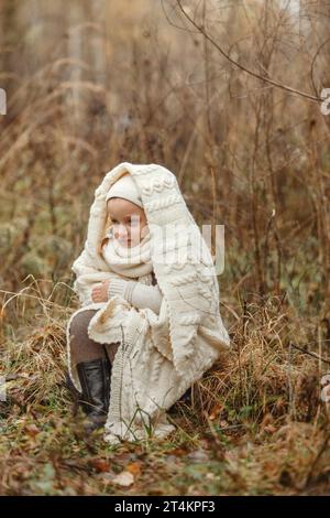 Portrait d'une jeune fille souriante assise dans un parc enveloppée dans une couverture blanche. Cadre vertical. Banque D'Images