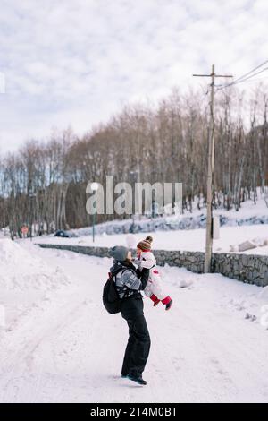 Maman lève haut dans ses bras un petit enfant sur une route enneigée dans le parc. Vue latérale Banque D'Images