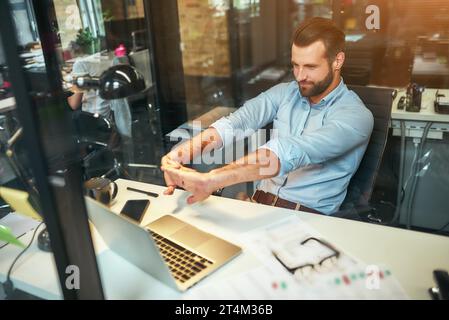 Détente sur le lieu de travail. Fatigué jeune homme d'affaires barbu en tenue formelle étirant ses bras et souriant tout en étant assis dans le bureau moderne Banque D'Images