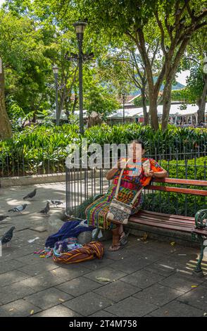 Guatemala, la Antigua - 20 juillet 2023 : Femme dans des tissus colorés traditionnels assis sur un banc sur la place centrale, plaza Mayor, vend de faux cou en plastique Banque D'Images