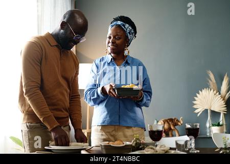 Heureuse jeune femme afro-américaine avec plateau de maïs cuit parlant à son mari en mettant des assiettes sur la table de fête tout en aidant sa femme Banque D'Images