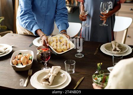 Cliché recadré de jeune femme mettant le plateau avec du maïs cuit au four sur la table de fête servi avec de la nourriture et des boissons maison pour le dîner de Thanksgiving Banque D'Images