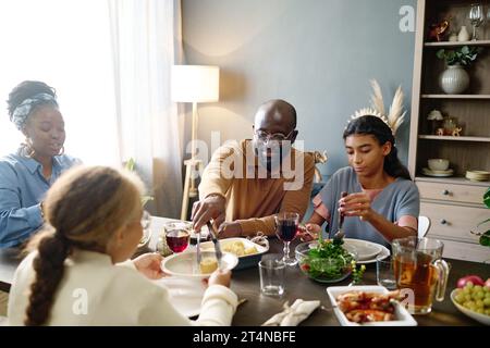 Jeune homme mettant du maïs sur l'assiette de sa fille sur la table de fête servie avec des boissons et de la nourriture maison tout en étant assis parmi les membres de la famille Banque D'Images