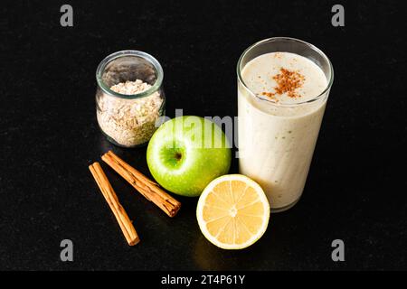 Un verre avec smoothie maison à côté d'un demi citron, une pomme verte, un pot avec des flocons d'avoine et deux bâtons de cannelle sur un banc de cuisine noir vu de Banque D'Images