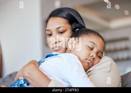 Jeune mère de race mixte regardant inquiet tout en tenant sa fille pendant qu'elle dort et assise sur le canapé dans le salon à la maison. Petite fille Banque D'Images