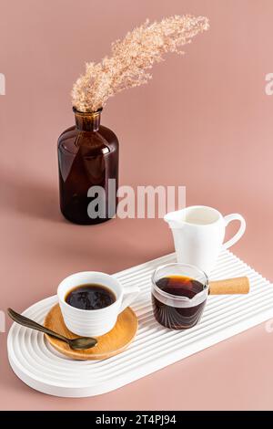 Une petite tasse de café espresso fraîchement préparé sur un plateau en céramique blanche en forme d'arc. un vase avec des branches sèches. Vue verticale Banque D'Images