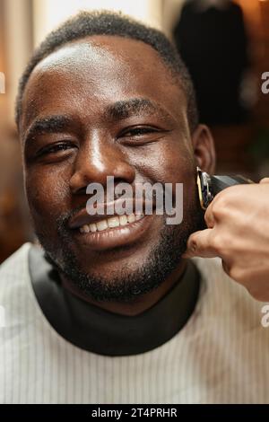 Portrait vertical de client afro-américain souriant à la caméra tout en obtenant la procédure de rasage dans le salon de coiffure Banque D'Images