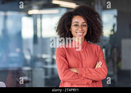Portrait heureuse femme d'affaires réussie, patron en chemise, souriant et regardant la caméra, bureau avec les bras croisés, femme afro-américaine avec les cheveux bouclés sur le bureau, programmeur informatique dans les lunettes. Banque D'Images