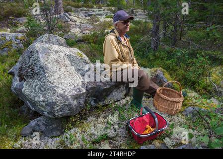 Femme âgée est assise sur un rocher dans la forêt et prend une courte pause après avoir cherché des chanterelles jaunes. Banque D'Images