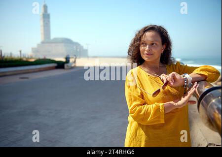 Belle femme multiethnique aux cheveux bouclés, tenant des lunettes de soleil, souriant regardant de côté, debout contre la mosquée Hassan II à Casablanca, Maroc. pe Banque D'Images