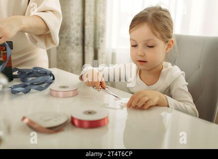 Belle petite fille prépare des cadeaux de Noël pour des amis et les emballe dans des boîtes tout en étant assis à la table Banque D'Images