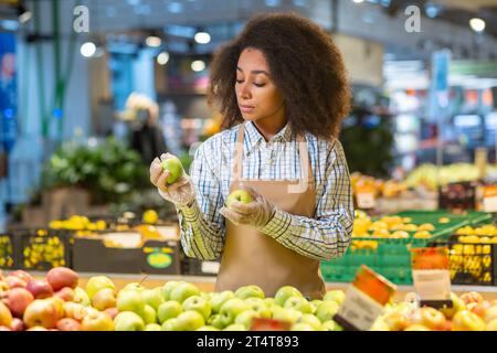 Travailleur de magasin de vendeur de femme concentrée sérieuse inspecte trie et trie les pommes dans le supermarché, parmi les fruits et légumes dans le département d'épicerie. Banque D'Images