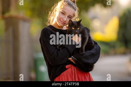 Jolie fille embrassant chat noir à l'extérieur dans la rue avec la lumière d'automne. Beau modèle adolescent tenant l'animal félin au parc Banque D'Images