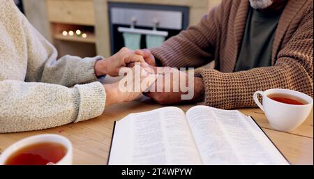 Étude de la Bible, couple senior et se tenant la main pour la prière, la religion et le gros plan à la maison pour la pleine conscience. Personne âgée, livre et espoir de paix Banque D'Images