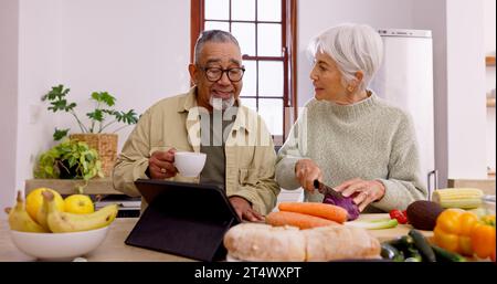 Couple senior, cuisine et tablette dans la cuisine à la maison avec couteau, légumes et tasse de thé pour la conversation. Mariage interracial, femme âgée et vieil homme Banque D'Images