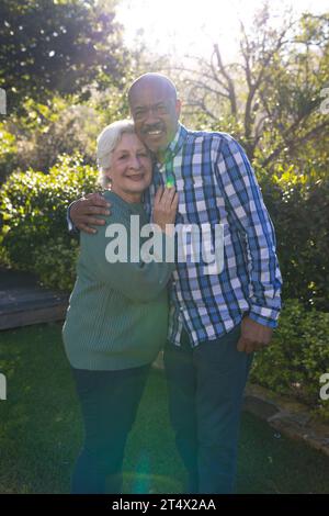 Heureux couple diversifié d'amis seniors embrassant et souriant dans le jardin ensoleillé Banque D'Images