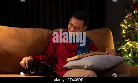 Un homme asiatique heureux dans un pull confortable et une écharpe caresse son chat mignon tout en lisant un livre et se relaxant sur un canapé dans le salon la nuit. Lifest Banque D'Images
