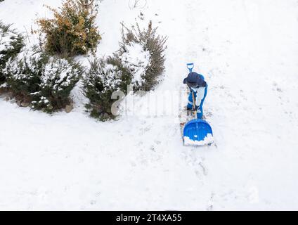 garçon d'environ 7 ans dans un costume d'hiver bleu chaud avec une grande pelle aide à dégager la neige dans la cour après une tempête de neige. Hiver temps de neige, mauvais temps Banque D'Images