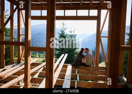 Homme et femme inspectant leur future maison de cadre en bois nichée dans les montagnes près de la forêt. Jeune couple sur le chantier en début de matinée. Concept de construction écologique contemporaine. Banque D'Images