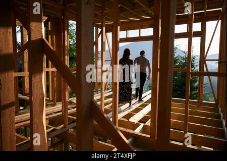 Homme et femme inspectant leur future maison de cadre en bois nichée dans les montagnes près de la forêt. Jeune couple sur le chantier en début de matinée. Concept de construction écologique contemporaine. Banque D'Images