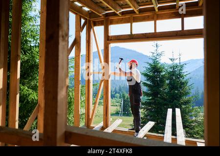 Menuisier construisant un cadre en bois, maison de deux étages près de la forêt. Homme barbu martelant des clous avec un marteau tout en portant un casque de protection et une combinaison de travail. Concept de construction écologique moderne. Banque D'Images