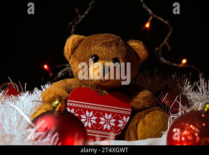 Un ours en peluche brun est assis avec un jouet d'arbre de Noël sous la forme d'un coeur sur un guirlard blanc, entouré de décorations rouges d'arbre de Noël. Dans le backgro Banque D'Images