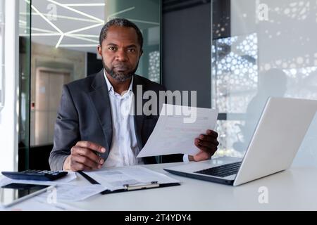 Portrait de mature strict homme d'affaires financier sérieux, homme regardant la caméra avec concentration et regard sérieux à la caméra, patron en costume d'affaires derrière le travail de papier à l'aide d'un ordinateur portable à l'intérieur du bureau. Banque D'Images