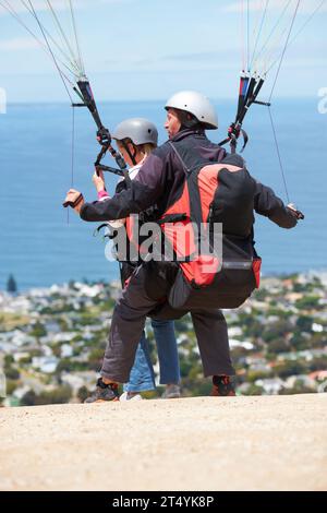 Les gens, le parapente et en plein air pour atterrir sur terre, le sport extrême et le soleil par l'océan sur la colline. Coach, personne et retour à l'aventure avec casque Banque D'Images