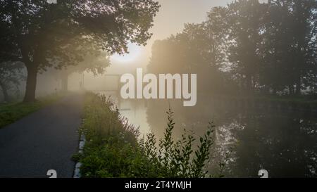 Dans les moments feutrés du petit matin, une scène d'une beauté inégalée se déroule. Le soleil levant, émergeant coyly de l'étreinte du brouillard, peint le ciel avec des teintes d'or et d'ambre. En dessous, les eaux calmes du canal agissent comme une toile, reflétant ce spectacle céleste. Sentinelle debout le long du bord de l'eau, les arbres jettent des silhouettes énigmatiques, ajoutant profondeur et mystère à ce paysage envoûtant. C'est l'orchestre de la nature qui joue une symphonie de lumière, d'ombre et de réflexion. Sunrise Serenade : mystique de brouillard, reflets et silhouettes d'arbre. Photo de haute qualité Banque D'Images