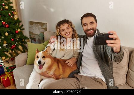 Couple heureux souriant et prenant la photo à l'appareil photo avec chien corgi près de l'arbre de Noël décoré Banque D'Images